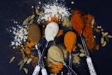 Five spoons hold different colored spices, surrounded by scattered powders and cardamom pods on a dark surface. The spices range from brown and white to orange and red, creating a colorful and textured composition.