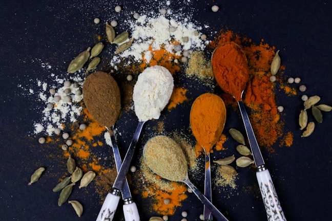 Five spoons hold different colored spices, surrounded by scattered powders and cardamom pods on a dark surface. The spices range from brown and white to orange and red, creating a colorful and textured composition.