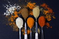 A dark background with five spoons of various spices arranged in a row. The different spices include a white powder, a brown powder, a yellow powder, an orange powder, and a red powder. Some spices are scattered around the spoons, creating a colorful and textured presentation.