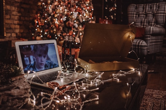 A cozy living room setting features a decorated Christmas tree with twinkling lights in the background. In the foreground, a laptop displays a movie, and next to it lies an open pizza box and a couple of soda bottles. String lights are casually draped across the table, adding to the festive atmosphere. A comfortable armchair with a plaid pattern adds to the homely feel.