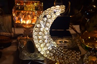A decorative crescent moon made of numerous circular crystals is prominently featured. Surrounding it are various ornate metallic objects, including an intricately designed lamp with a warm glow and a metal box with floral engravings. The scene conveys an atmosphere of luxury and elegance.