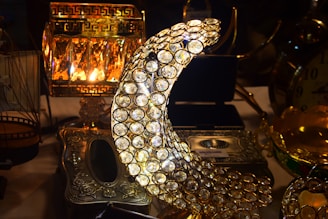 A decorative crescent moon made of numerous circular crystals is prominently featured. Surrounding it are various ornate metallic objects, including an intricately designed lamp with a warm glow and a metal box with floral engravings. The scene conveys an atmosphere of luxury and elegance.