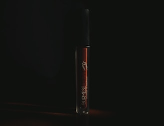 A tube of liquid lipstick stands upright against a dark background. The branding on the tube reads 'Burmese Dream', indicating the product name. The lighting is low, casting a moody atmosphere around the tube, which has a sleek, glossy appearance.
