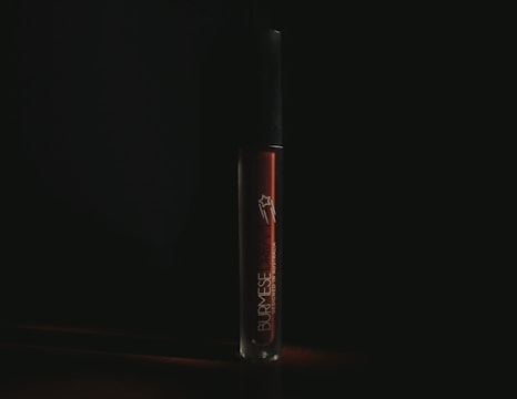 A tube of liquid lipstick stands upright against a dark background. The branding on the tube reads 'Burmese Dream', indicating the product name. The lighting is low, casting a moody atmosphere around the tube, which has a sleek, glossy appearance.