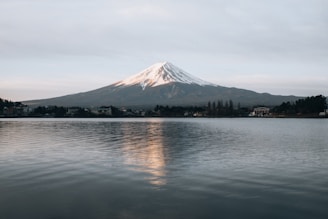 Mount Fuji stands majestically with a snow-capped peak under a slightly cloudy sky. The calm waters of the lake in the foreground reflect the mountain and the surrounding landscape, including sparse trees and buildings near the shore.