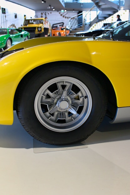 A close-up view of a vintage car wheel with a shiny metallic hubcap and a black tire. The car's body is yellow, and there are additional cars in the background with various colors such as green and orange, displayed in a modern showroom with sleek designs and a staircase.