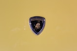 A Lamborghini emblem is prominently displayed against a yellow background. The emblem features a black shield with a golden bull in the center and the brand name written above.