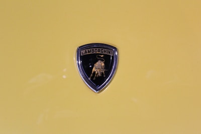 A Lamborghini emblem is prominently displayed against a yellow background. The emblem features a black shield with a golden bull in the center and the brand name written above.