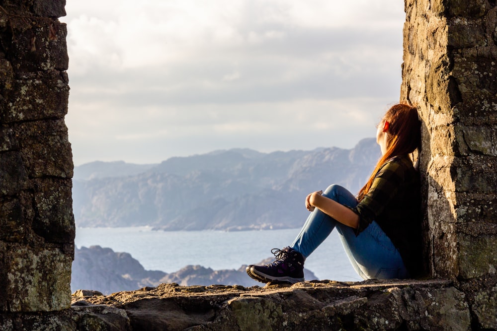 woman in black jacket sitting on rock near body of water during daytime