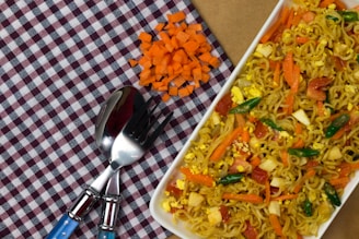 A white rectangular plate filled with stir-fried noodles mixed with chopped vegetables such as carrots, bell peppers, and small pieces of egg is placed on a checkered cloth. Beside it, there is a small pile of neatly chopped orange carrots. A fork and a spoon with blue handles are placed on the cloth near the noodles.