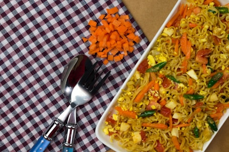 A white rectangular plate filled with stir-fried noodles mixed with chopped vegetables such as carrots, bell peppers, and small pieces of egg is placed on a checkered cloth. Beside it, there is a small pile of neatly chopped orange carrots. A fork and a spoon with blue handles are placed on the cloth near the noodles.