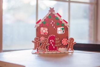 A festive gingerbread house decorated with various candy-themed ornaments sits on a table. The house features red and white peppermint swirls, green gumdrop shapes, and candy canes. Two gingerbread figures stand around it, framed by a soft focus window in the background allowing natural light to illuminate the scene.