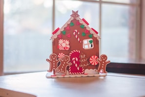 A festive gingerbread house decorated with various candy-themed ornaments sits on a table. The house features red and white peppermint swirls, green gumdrop shapes, and candy canes. Two gingerbread figures stand around it, framed by a soft focus window in the background allowing natural light to illuminate the scene.
