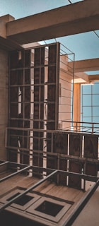 A partially constructed building with visible framework and metal railings. The structure includes open spaces for windows and exposed beams, highlighting the unfinished state. The lighting casts shadows creating a contrast with the brighter blue sky visible through openings.