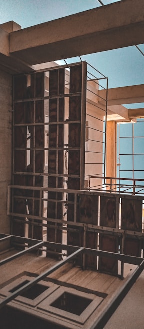 A partially constructed building with visible framework and metal railings. The structure includes open spaces for windows and exposed beams, highlighting the unfinished state. The lighting casts shadows creating a contrast with the brighter blue sky visible through openings.