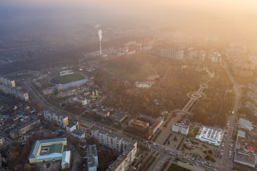 An aerial view of an urban area with a mix of buildings, a smoke stack emitting smoke, large parks with circular designs and pathways, and several roads and intersections. The image has a hazy atmosphere, possibly due to fog or pollution, with the sky taking on a gradient of light yellow-orange hues as the light diffuses.