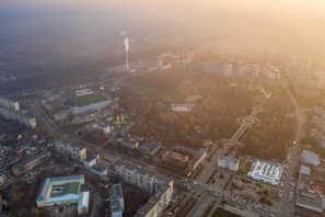 An aerial view of an urban area with a mix of buildings, a smoke stack emitting smoke, large parks with circular designs and pathways, and several roads and intersections. The image has a hazy atmosphere, possibly due to fog or pollution, with the sky taking on a gradient of light yellow-orange hues as the light diffuses.