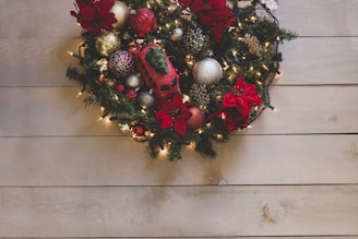 A decorative Christmas wreath adorned with red and gold baubles, pinecones, and poinsettias. It is enhanced by small string lights that add a warm glow. The wreath is placed against a light wooden background, creating a festive holiday appearance.