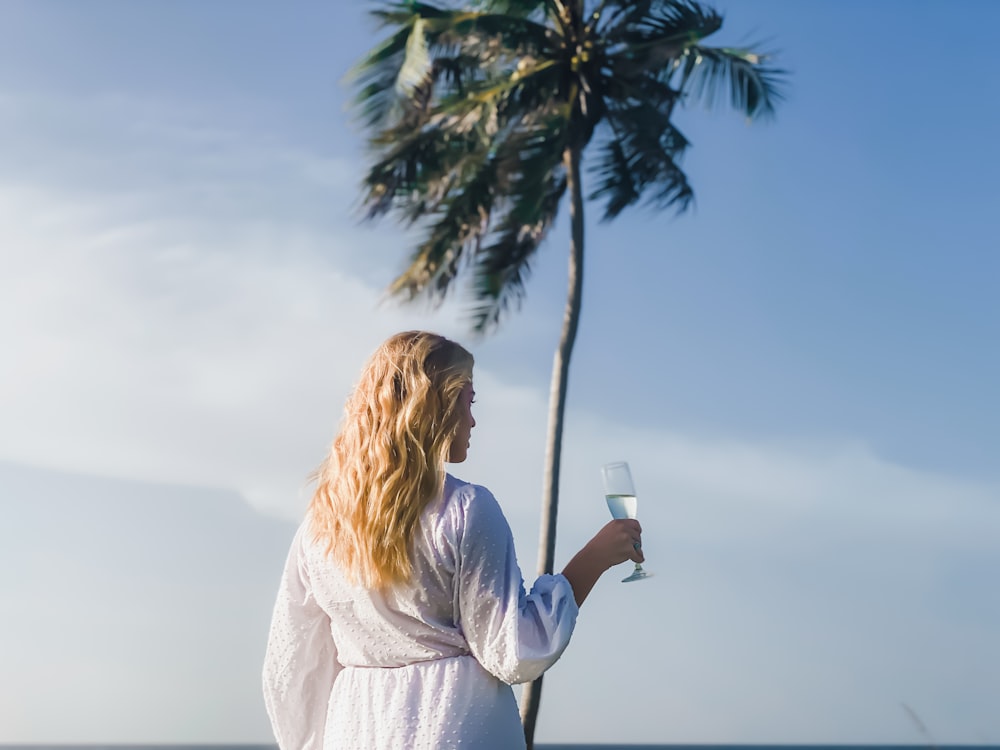 donna in camicia bianca a maniche lunghe che tiene tazza di ceramica bianca in piedi accanto alla palma durante il giorno