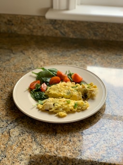 A plate with a portion of scrambled eggs, accompanied by fresh spinach leaves and sliced cherry tomatoes, placed on a granite countertop near a sunlit window.