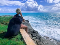 woman in black dress sitting on gray concrete bench near body of water during daytime