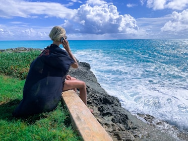 woman in black dress sitting on gray concrete bench near body of water during daytime