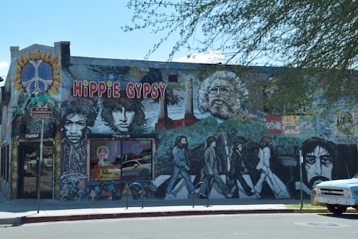 A mural on the side of a building features vibrant portraits and a peace sign. The artwork includes iconic figures and psychedelic elements, accompanied by the text 'Hippie Gypsy' in bold letters. Bright colors and pop culture references create a lively and artistic atmosphere.