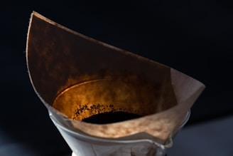 A close-up view of a coffee filter filled with dark coffee grounds, set inside a glass dripper. The lighting emphasizes the texture and color of the used coffee filter, casting warm tones against a dark background.