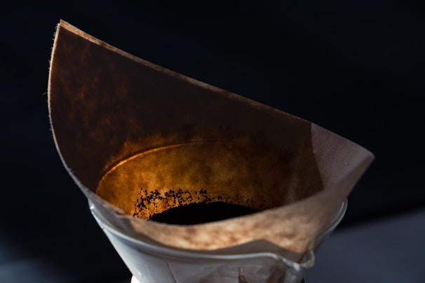 A close-up view of a coffee filter filled with dark coffee grounds, set inside a glass dripper. The lighting emphasizes the texture and color of the used coffee filter, casting warm tones against a dark background.