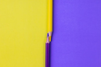 Two colored pencils are placed against a split background, with the yellow pencil aligned to the yellow side and the purple pencil aligned to the purple side. The pencils are positioned vertically and meet at the middle, creating a point of focus at their tips.