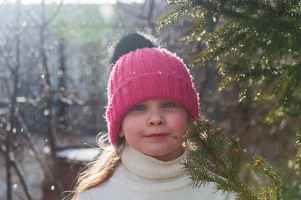girl in white turtleneck sweater and red knit cap standing beside green pine tree during daytime