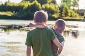 Two children are standing near a serene lake, with one arm around the other's shoulder. The lush greenery and trees, along with the reflective water, create a peaceful outdoor setting. The sunlight casts a warm glow over the scene, emphasizing the bond between the two individuals.