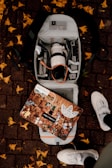 An open backpack on a brick sidewalk reveals a camera lens, various electronic devices, and accessories. A decorated laptop with multiple stickers depicting various characters and logos rests on top of the backpack. Yellow leaves are scattered around on the ground, and a pair of white sneakers is visible at the bottom of the image.