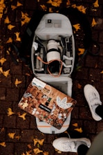 An open backpack on a brick sidewalk reveals a camera lens, various electronic devices, and accessories. A decorated laptop with multiple stickers depicting various characters and logos rests on top of the backpack. Yellow leaves are scattered around on the ground, and a pair of white sneakers is visible at the bottom of the image.