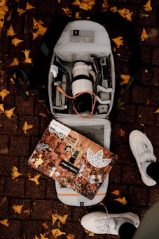 An open backpack on a brick sidewalk reveals a camera lens, various electronic devices, and accessories. A decorated laptop with multiple stickers depicting various characters and logos rests on top of the backpack. Yellow leaves are scattered around on the ground, and a pair of white sneakers is visible at the bottom of the image.