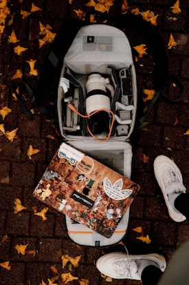 An open backpack on a brick sidewalk reveals a camera lens, various electronic devices, and accessories. A decorated laptop with multiple stickers depicting various characters and logos rests on top of the backpack. Yellow leaves are scattered around on the ground, and a pair of white sneakers is visible at the bottom of the image.