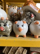 Several ceramic piggy banks are arranged closely together on a shelf. The piggy banks are primarily white with orange accents on their feet, ears, and snouts. Some have black, marble-like patterns on them. The shelf is yellow, and there are various other items visible in the background.