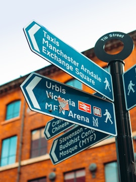 Several blue street signs displaying directions to various locations like Taxis, Manchester Arndale, and Exchange Square against a background of a red brick building. The signs are mounted on a black pole at an intersection.