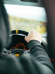 A person is holding a steering wheel inside a car, with a focus on the dashboard displaying illuminated gauges. The steering wheel has branding marked 'OMP', and the view through the windshield suggests motion.