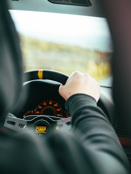 A person is holding a steering wheel inside a car, with a focus on the dashboard displaying illuminated gauges. The steering wheel has branding marked 'OMP', and the view through the windshield suggests motion.