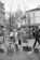 A black and white photo depicts an outdoor art market. Several easels display paintings, with subjects including portraits and architecture. People dressed in coats are engaged in viewing or discussing the artworks. In the background, the iconic dome of a large building is visible past leafless trees and surrounding architecture.