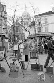 A black and white photo depicts an outdoor art market. Several easels display paintings, with subjects including portraits and architecture. People dressed in coats are engaged in viewing or discussing the artworks. In the background, the iconic dome of a large building is visible past leafless trees and surrounding architecture.