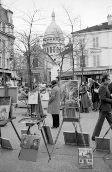 A black and white photo depicts an outdoor art market. Several easels display paintings, with subjects including portraits and architecture. People dressed in coats are engaged in viewing or discussing the artworks. In the background, the iconic dome of a large building is visible past leafless trees and surrounding architecture.
