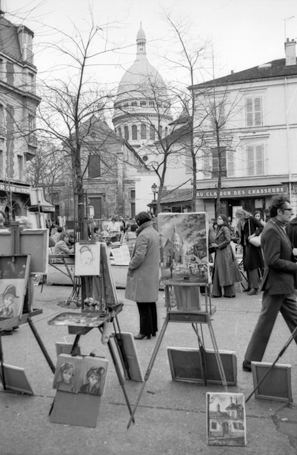 A black and white photo depicts an outdoor art market. Several easels display paintings, with subjects including portraits and architecture. People dressed in coats are engaged in viewing or discussing the artworks. In the background, the iconic dome of a large building is visible past leafless trees and surrounding architecture.