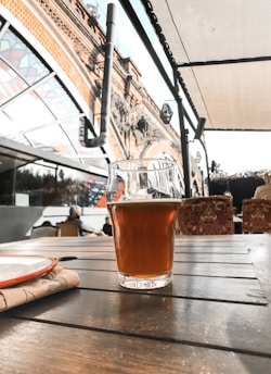 A glass of amber-colored beer sits on a wooden table in an outdoor seating area. The background features an ornate, arched brick structure and a stained glass canopy, with seating furniture and people visible.