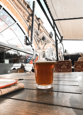 A glass of amber-colored beer sits on a wooden table in an outdoor seating area. The background features an ornate, arched brick structure and a stained glass canopy, with seating furniture and people visible.