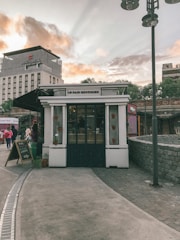 A small café named 'Le Pain Quotidien' is located along a paved path, with potted plants hanging on the walls. A chalkboard sign stands outside. In the background, a multi-story building displays the name 'SWISS MEDICAL'. The sky is filled with clouds, suggesting either sunrise or sunset.
