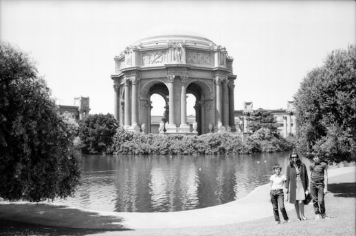 An ornate, classical-style rotunda stands prominently by a reflective pond, surrounded by lush greenery. Three people are gathered near the water's edge, casting shadows on the grass.