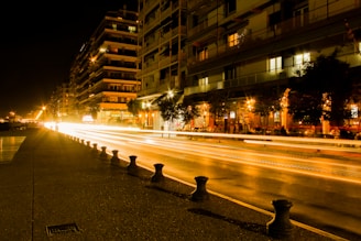 A nighttime urban scene features brightly lit buildings alongside a road with light trails indicating the movement of cars. The warm glow of the streetlights and the reflections create a lively atmosphere. People are seated at an outdoor café, adding to the bustling nightlife.