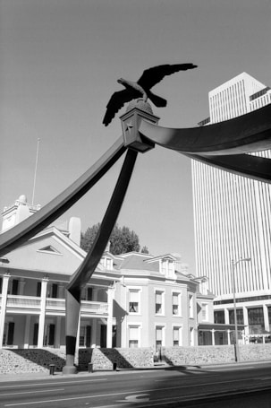 A monochrome photograph featuring a striking structural element with an eagle sculpture perched atop it. The scene includes a historic building with columned balconies, evoking a classical architectural style, adjacent to a modern high-rise office building. The contrast between the traditional and contemporary structures adds depth to the scene.